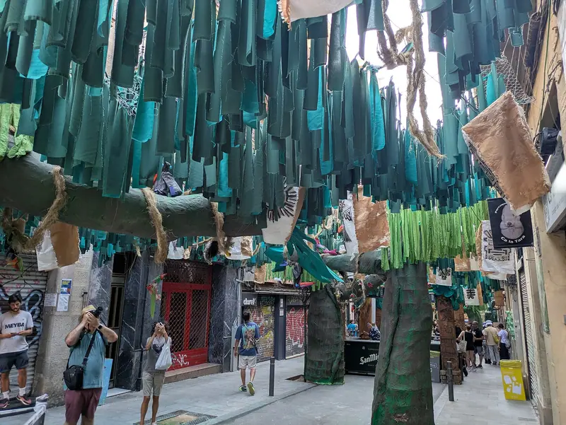 Calle de Gràcia decorada para la fiesta mayor con tiras de tela verde colgadas y troncos de papel, con gente mirando