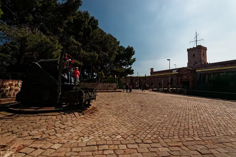Explanada empedrada junto al castillo de Montjuïc, con un cañón antiguo a la izquierda, pinos y algunos visitantes al fondo